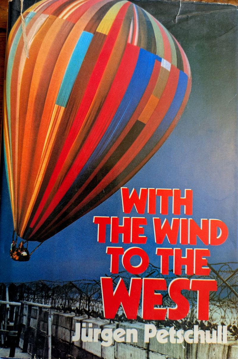 Picture of colour pictorial book cover showing colourful patchwork hot air balloon airborne in dark blue sky above barbed wire on top of Berlin wall